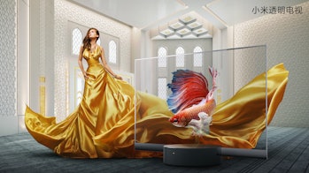 A model in a deep yellow silky dress is seen standing behind a Xiaomi see-through TV set. The background depicts grafting on a white wall and ornamental designs on the ceiling. There is a fish on the transparent screen.